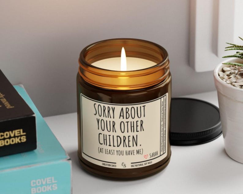May include: A lit soy wax candle in a brown glass jar with a black lid. The candle has a white label with the text "SORRY ABOUT YOUR OTHER CHILDREN. (AT LEAST YOU HAVE ME)." The candle is next to a small potted plant and books.
