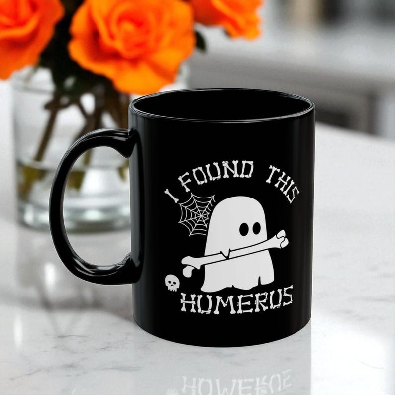 Op de afbeelding: Een zwarte keramische mok met een witte afbeelding van een geest die een bot vasthoudt. De tekst I FOUND THIS HUMERUS staat in een bot-achtig lettertype. Een klein spinnenweb en een schedel maken het griezelige ontwerp op de mok compleet.