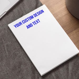 以下が含まれることがあります： 上部に青い文字で「YOUR CUSTOM DESIGN AND TEXT」と印刷された白いメモ帳です。
