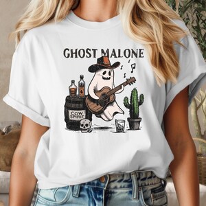 May include: White t-shirt with a graphic of a ghost wearing a cowboy hat and playing a guitar. The text "Ghost Malone" is printed above the ghost. The ghost is holding a bottle of "Cow Spirit" liquor.
