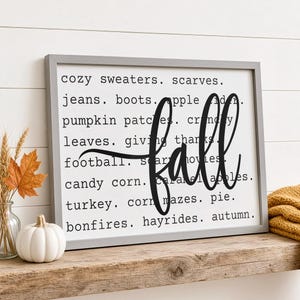 May include: A rectangular wall sign with a gray frame and white background featuring black text listing fall-themed words including cozy sweaters, scarves, jeans, boots, apple cider, pumpkin patches, crunchy leaves, giving thanks, football, scary movies, candy corn, caramel apples, turkey, corn mazes, pie, bonfires, hayrides, and autumn, with the word fall in large script.