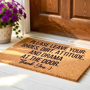 May include: A rectangular coir doormat with the message "PLEASE LEAVE YOUR: SHOES, DIRT, ATTITUDE, AND DRAMA AT THE DOOR. Thank You :)" in black. The mat is placed in front of a wooden door.