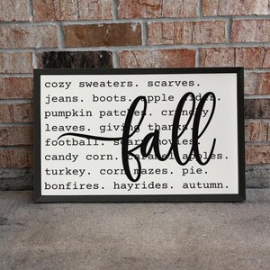 May include: A rectangular wall sign with a black frame and a white background featuring black text listing fall-themed words like sweaters, boots, and pumpkin patches, with the word fall written in a large, black, cursive script across the center.