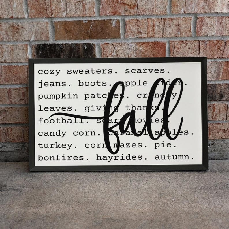 May include: A rectangular wall sign with a black frame and a white background featuring black text listing fall-themed words like sweaters, boots, and pumpkin patches, with the word fall written in a large, black, cursive script across the center.