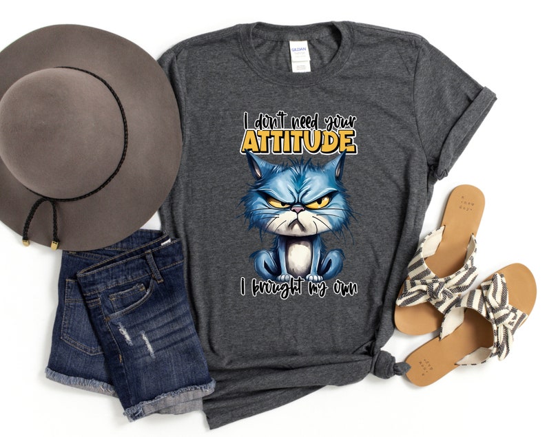 Op de afbeelding: Een donkergrijs T-shirt met een cartoonkat met een chagrijnige uitdrukking. De tekst op het shirt luidt "I don't need your attitude I brought my own".