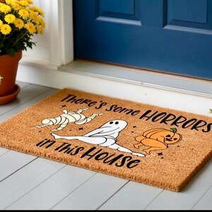 May include: A rectangular coir doormat with the text "There's some HORRORS in this HOUSE". The mat features illustrations of a mummy, a ghost, and a pumpkin. The mat is placed in front of a blue door.