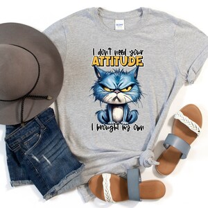 Op de afbeelding: Grijs T-shirt met een blauwe cartoonkat en de tekst "Ik heb je houding niet nodig, ik heb mijn eigen meegebracht".