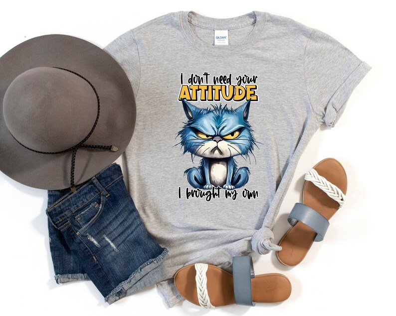 Op de afbeelding: Grijs T-shirt met een blauwe cartoonkat en de tekst "Ik heb je houding niet nodig, ik heb mijn eigen meegebracht".