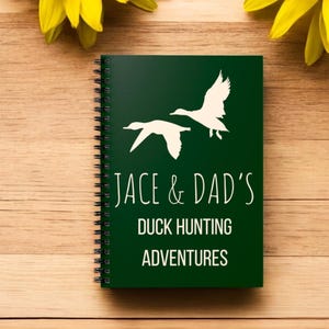 May include: A green spiral bound notebook with white silhouettes of ducks flying. The notebook has the text "Jace & Dad's Duck Hunting Adventures" printed on the front.