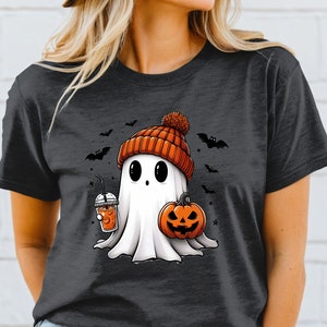 May include: A dark grey t-shirt featuring a white ghost wearing an orange knitted hat and holding a pumpkin and a cup of orange drink. The ghost has large eyes and a small mouth. There are bats and stars in the background.