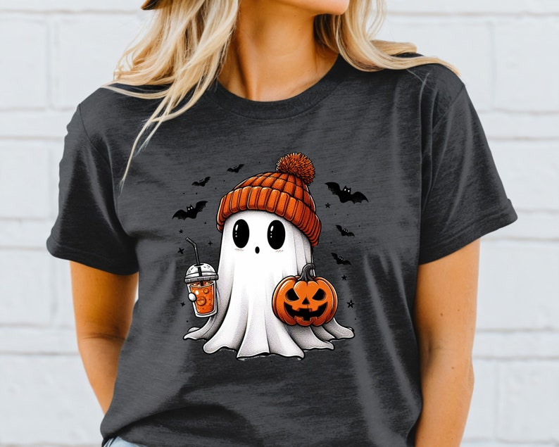 May include: A dark grey t-shirt featuring a white ghost wearing an orange knitted hat and holding a pumpkin and a cup of orange drink. The ghost has large eyes and a small mouth. There are bats and stars in the background.
