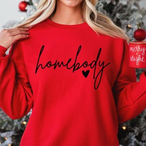 K&ouml;nnte beinhalten: Roter Pullover mit dem Text "homebody" in schwarzer Schrift mit einem kleinen Herzen. Die Person h&auml;lt eine rote Tasse mit dem Text "merry + bright" in wei&szlig;.