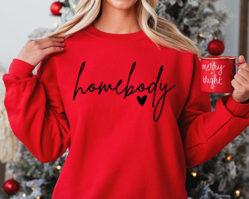 K&ouml;nnte beinhalten: Roter Pullover mit dem Text "homebody" in schwarzer Schrift mit einem kleinen Herzen. Die Person h&auml;lt eine rote Tasse mit dem Text "merry + bright" in wei&szlig;.
