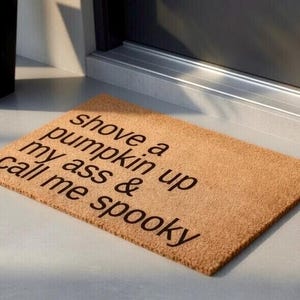 Op de afbeelding: Een rechthoekige deurmat van natuurlijke vezels. De mat is lichtbruin van kleur en heeft de tekst "shove a pumpkin up my ass & call me spooky" in donkerbruine letters. De mat ligt voor een deur.