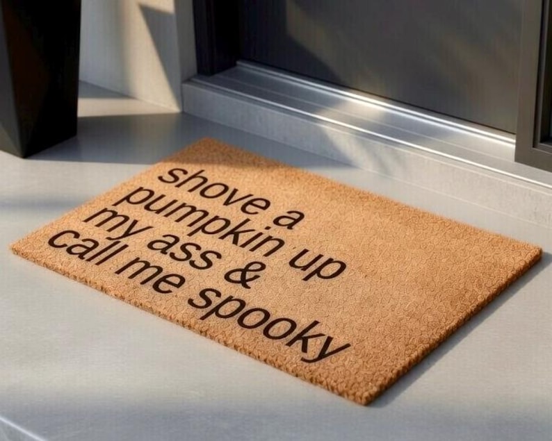 Op de afbeelding: Een rechthoekige deurmat van natuurlijke vezels. De mat is lichtbruin van kleur en heeft de tekst "shove a pumpkin up my ass & call me spooky" in donkerbruine letters. De mat ligt voor een deur.