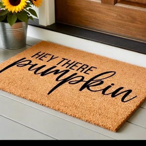 May include: A rectangular doormat made of natural coir fiber. The mat features the phrase "HEY THERE PUMPKIN" in a black, cursive font. The mat is placed on a gray painted wooden porch.