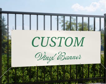 Puede incluir: Una pancarta de vinilo color crema con las palabras "CUSTOM" y "Vinyl Banner" en letras verdes. La pancarta está sujeta a una valla metálica negra con cuerdas blancas, sobre un fondo de follaje verde y cielo azul.
