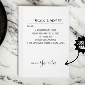 May include: A white notebook with a black and white definition of "Boss Lady" with a heart symbol. The definition reads: "A strong and intelligent woman who completes all task set before her with confidence and grace, a true super human and multitasking expert." The notebook also includes the text "See also: Jennifer" and a black starburst with the text "CUSTOM NAME".