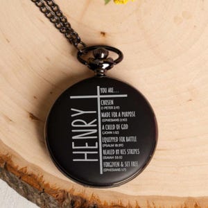 May include: Black pocket watch with a silver chain and a cross design. The watch face has the name "HENRY" engraved on it and a list of bible verses.
