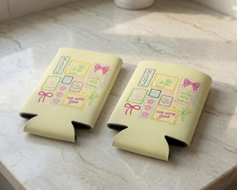 Puede incluir: Dos aislantes para bebidas de color amarillo p&aacute;lido con un estampado gr&aacute;fico colorido de fichas de Mahjong, lazos rosas, flores de cerezo y tallos de bamb&uacute; verdes. El texto "MAHJ" y "one more game" aparece entre los iconos.