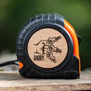 May include: A black and orange tape measure with a wooden face. The face has a design of four fists with the names "Daddy", "Layla", "Aiden", and "Harper" written on them.