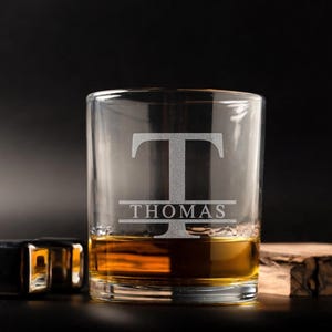 May include: A clear rocks glass with the name "THOMAS" and the letter "T" etched into the glass. The glass is filled with amber liquid. Two stainless steel ice cubes and a piece of wood are next to the glass.