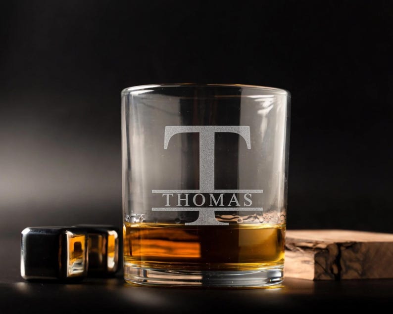 May include: A clear rocks glass with the name "THOMAS" and the letter "T" etched into the glass. The glass is filled with amber liquid. Two stainless steel ice cubes and a piece of wood are next to the glass.