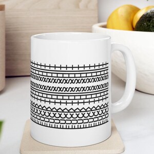 K&ouml;nnte beinhalten: Wei&szlig;e Keramik-Kaffeetasse mit einem schwarzen geometrischen Muster. Das Design zeigt horizontale Linien, Zickzackmuster und gebogene Formen. Die Tasse hat einen Standardgriff und wird wahrscheinlich f&uuml;r Hei&szlig;getr&auml;nke verwendet. Die Tasse steht auf einem beigen Untersetzer.
