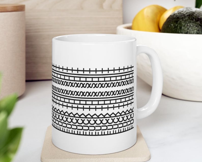 K&ouml;nnte beinhalten: Wei&szlig;e Keramik-Kaffeetasse mit einem schwarzen geometrischen Muster. Das Design zeigt horizontale Linien, Zickzackmuster und gebogene Formen. Die Tasse hat einen Standardgriff und wird wahrscheinlich f&uuml;r Hei&szlig;getr&auml;nke verwendet. Die Tasse steht auf einem beigen Untersetzer.