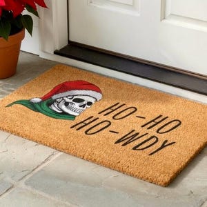 May include: A rectangular doormat with a skull wearing a Santa hat and green scarf. The text "HO-HO HO-WDY" is printed in black. The mat is made of a natural fiber material and is placed in front of a white door.