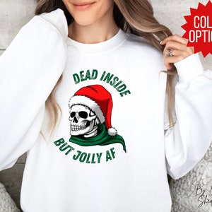 May include: White sweatshirt with a green and red graphic of a skull wearing a Santa hat. The text on the sweatshirt reads "Dead Inside But Jolly AF".