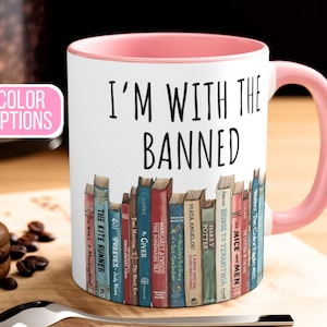 May include: A white ceramic mug with a pink rim and handle. The mug features a black and white graphic of a bookshelf with a variety of books. The text "I'm with the Banned" is printed in black on the mug.