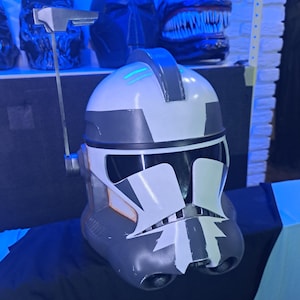 May include: A white and grey helmet with a black visor and accents, resembling a clone trooper helmet. The helmet features a distinctive design with a visor and a top antenna. Other helmets are visible in the background.