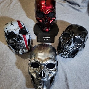 K&ouml;nnte beinhalten: Vier Totenkopfmasken in verschiedenen Metallic-Ausf&uuml;hrungen. Eine ist rot, auf einem St&auml;nder mit einem Namensschild "Michael Crist" montiert. Weitere Masken sind silber, schwarz und wei&szlig; mit roten Akzenten. Alle haben Augen- und Nasen&ouml;ffnungen.