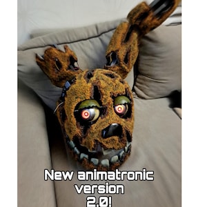 May include: A detailed, brown animatronic mask with glowing red eyes and a wide, toothy grin. The mask has a textured, moss-like appearance and the text "New animatronic version 2.0!" is displayed at the bottom.