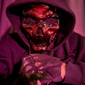 K&ouml;nnte beinhalten: Eine metallisch rote Totenkopfmaske mit aufwendigen Details, getragen mit einem schwarzen Kapuzenpullover und schwarzen Handschuhen. Die Maske hat eine gl&auml;nzende Oberfl&auml;che und ein skelettartiges Aussehen. Die Handschuhe haben ein strukturiertes Design.