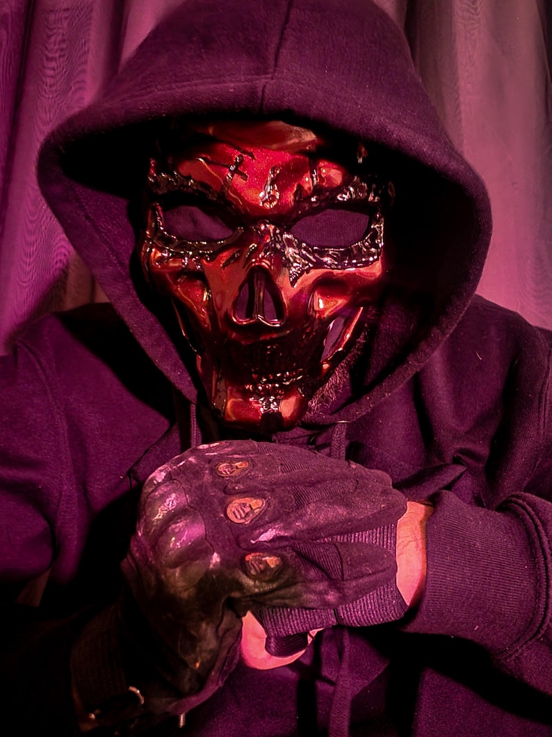 K&ouml;nnte beinhalten: Eine metallisch rote Totenkopfmaske mit aufwendigen Details, getragen mit einem schwarzen Kapuzenpullover und schwarzen Handschuhen. Die Maske hat eine gl&auml;nzende Oberfl&auml;che und ein skelettartiges Aussehen. Die Handschuhe haben ein strukturiertes Design.