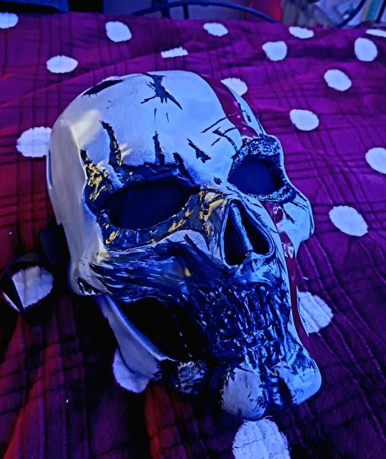 K&ouml;nnte beinhalten: Eine wei&szlig;-schwarze Totenkopfmaske mit einem Used-Design. Die Maske hat gro&szlig;e Augenh&ouml;hlen und einen detaillierten Kiefer. Die Maske liegt auf einem lila Stoff mit wei&szlig;en Punkten.