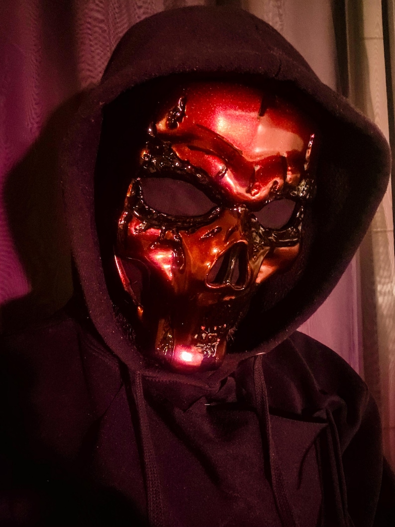 K&ouml;nnte beinhalten: Eine metallisch rote Totenkopfmaske mit aufwendigen Details, getragen mit einem schwarzen Kapuzenpullover. Die Maske hat eine gl&auml;nzende Oberfl&auml;che und Augen- und Nasen&ouml;ffnungen. Die Gesamt&auml;sthetik ist d&uuml;ster und geheimnisvoll.