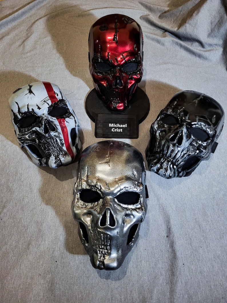 K&ouml;nnte beinhalten: Vier Totenkopfmasken in verschiedenen Metallic-Ausf&uuml;hrungen. Eine ist rot, auf einem St&auml;nder mit einem Namensschild "Michael Crist" montiert. Weitere Masken sind silber, schwarz und wei&szlig; mit roten Akzenten. Alle haben Augen- und Nasen&ouml;ffnungen.