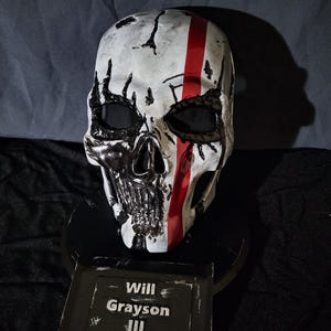 K&ouml;nnte beinhalten: Eine wei&szlig;e Totenkopfmaske mit schwarzen Details und einem roten Streifen. Die Maske ist auf einem schwarzen Sockel mit einem Namensschild mit der Aufschrift "Will Grayson III" montiert.