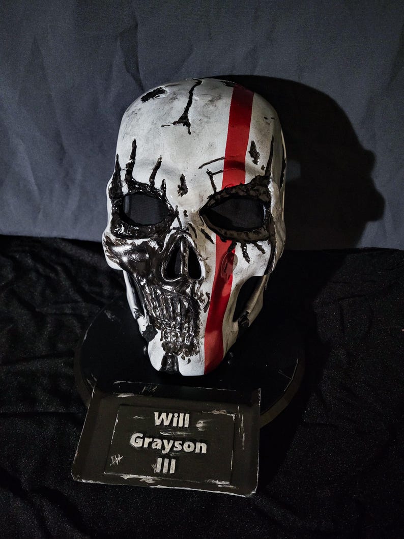 K&ouml;nnte beinhalten: Eine wei&szlig;e Totenkopfmaske mit schwarzen Details und einem roten Streifen. Die Maske ist auf einem schwarzen Sockel mit einem Namensschild mit der Aufschrift "Will Grayson III" montiert.