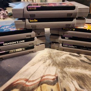 May include: Stacks of gray Super Nintendo video game cartridges. Titles visible include "Fatal Fury", "Dragon's Lair", "Yoshi's Cookie", and "Secret of Mana". The cartridges are stacked on a patterned surface.