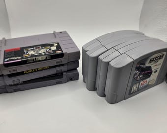 May include: A collection of gray plastic video game cartridges for a home console. One stack of three cartridges features labels with text including Williams Greatest Hits, Fighter's History, and Fatal Fury. A second stack features NASCAR 99.