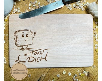 May include: A wooden cutting board with a cartoon drawing of a slice of bread with a face and the text "auf ein Toast auf Dich". The text "Auf Wunsch personalisiert" is also visible on the board.