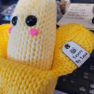 Puede incluir: Un juguete de peluche de plátano tejido con una cáscara amarilla y un plátano blanco en el interior. El plátano tiene ojos negros y mejillas rosadas. El juguete está sentado sobre una superficie de tejido amarillo. El juguete tiene una etiqueta que dice "Layers by Layla".