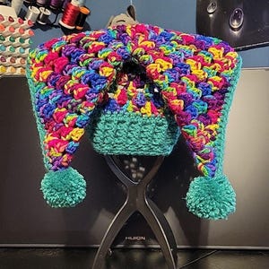 May include: A multi-colored crochet hat featuring a vibrant mix of pink, blue, yellow, and purple yarn. The hat has a solid teal brim and two long, dangling side flaps, each finished with a matching teal yarn pom-pom at the bottom end.
