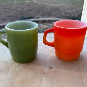 Puede incluir: Dos tazas de vidrio vintage, una verde y otra naranja. Ambas tienen asa y diseño apilado. Las tazas están sobre una superficie de madera. Perfectas para café o té.