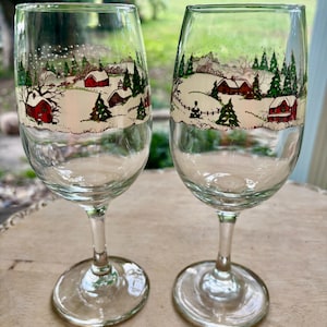 May include: Two clear wine glasses with a winter scene design. The design features red houses, green trees, and a snowy landscape. The glasses have a stem and a round base.
