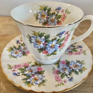 May include: A white teacup and saucer set with a floral pattern. The cup and saucer are decorated with blue and pink flowers on a white background. The cup has a gold rim and the saucer has a gold rim and a scalloped edge.
